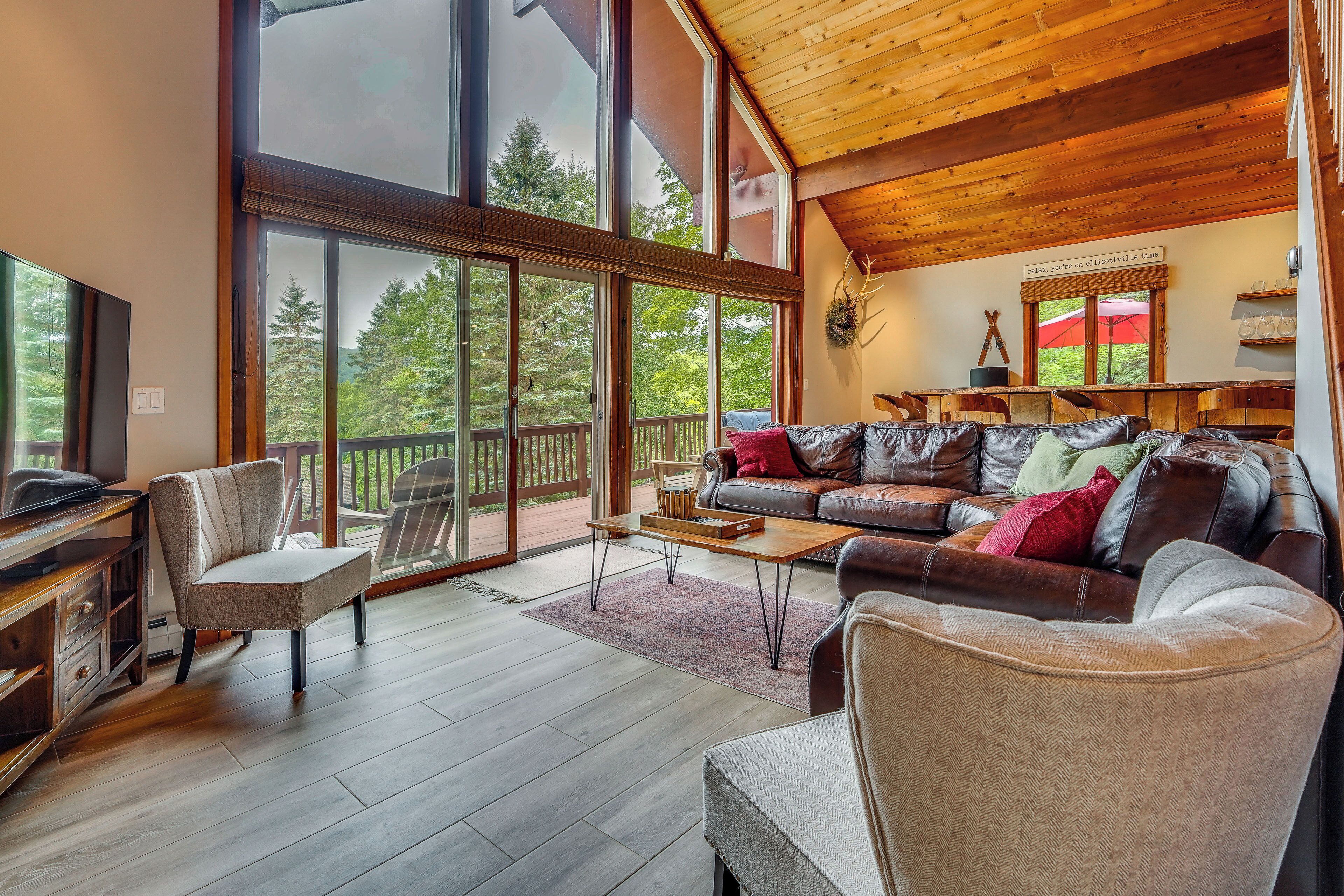 A spacious living area with a large wooden ceiling, a leather sofa, and a view of a forest through large windows.