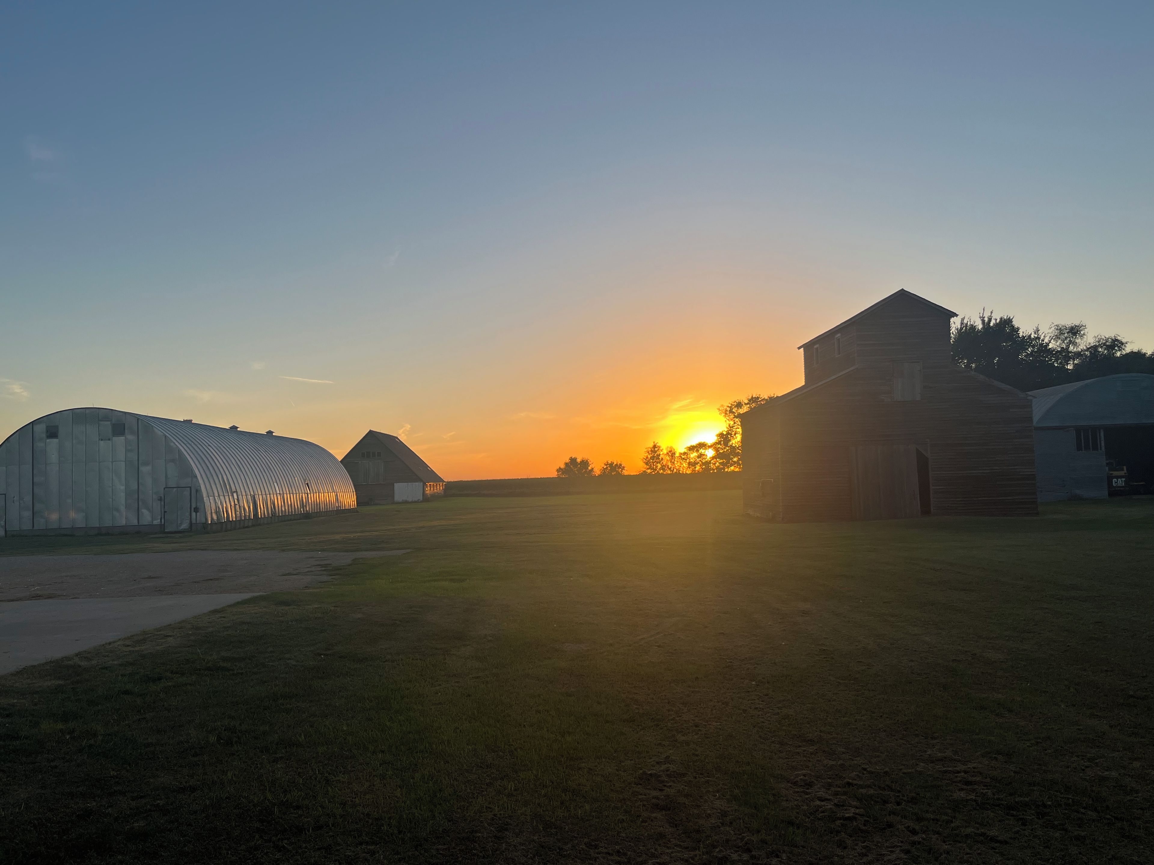 A rural landscape with a barn and a greenhouse during sunset.