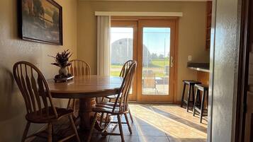A dining area with a wooden table and chairs, a painting on the wall, and a view through a glass door to the outside.