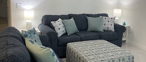 A living room with a dark-colored sofa set, a matching ottoman, and a wall-mounted picture.