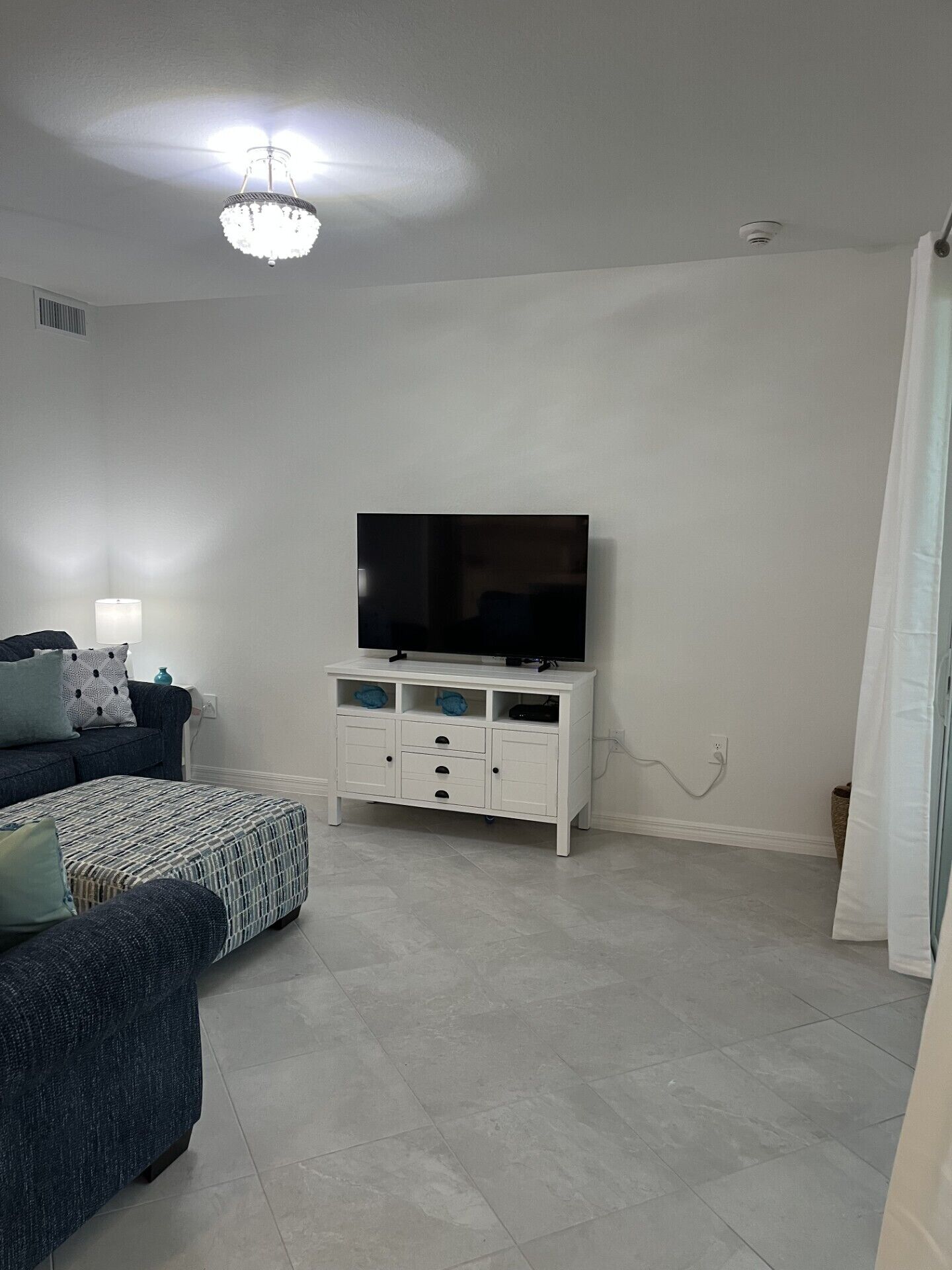 Una sala de estar con una lámpara en el techo, un televisor de pantalla plana sobre un mueble blanco, un sofá y un reposapiés estampado.