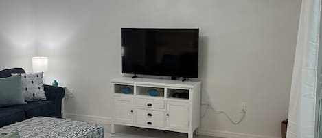 Una sala de estar con una lámpara en el techo, un televisor de pantalla plana sobre un mueble blanco, un sofá y un reposapiés estampado.
