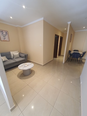 A living room with a sofa, a coffee table, and a dining area with chairs and a table. - Opportunité immobilière  (Hydra)