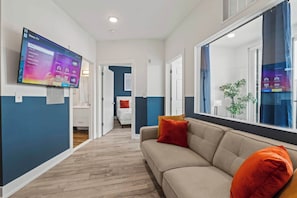 Ein modernes Hotelzimmer mit einem großen Flachbildfernseher an der Wand, einer Couch mit orangefarbenen Kissen und einem großen Fenster mit Ausblick.