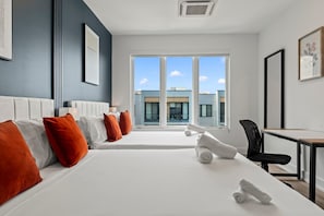Ein Hotelzimmer mit zwei Betten, einem Schreibtisch und Blick auf Gebäude durch große Fenster.