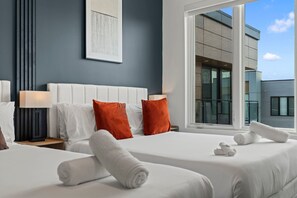 A hotel room with two beds, white linens, orange pillows, a large window, and a view of a building. - Modern 2BR | Sleeps 6 | Northern Liberties (Philadelphia)