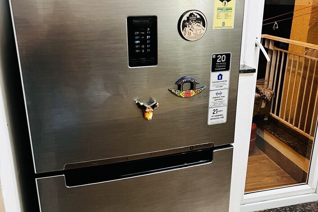 A stainless steel refrigerator with a digital display, a 'Twin Cooling' label, a 'Kashmir' decorative magnet, a '20' compressor label, and a '21 years of reliability' badge.