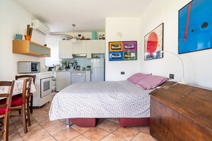 A compact living space with a kitchenette, a bed with a floral bedspread, a wooden dresser, and wall art.
