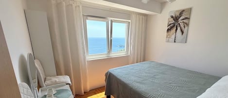 A hotel room with a bed, a chair with a tray, a window with curtains, and a painting of a palm tree.