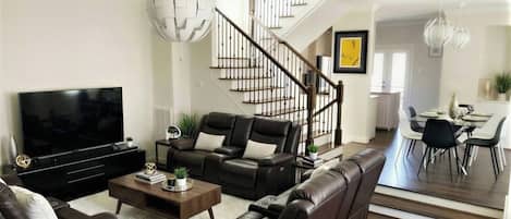 A modern living room with a flat-screen TV, leather sofas, a coffee table, and a staircase leading to a dining area.