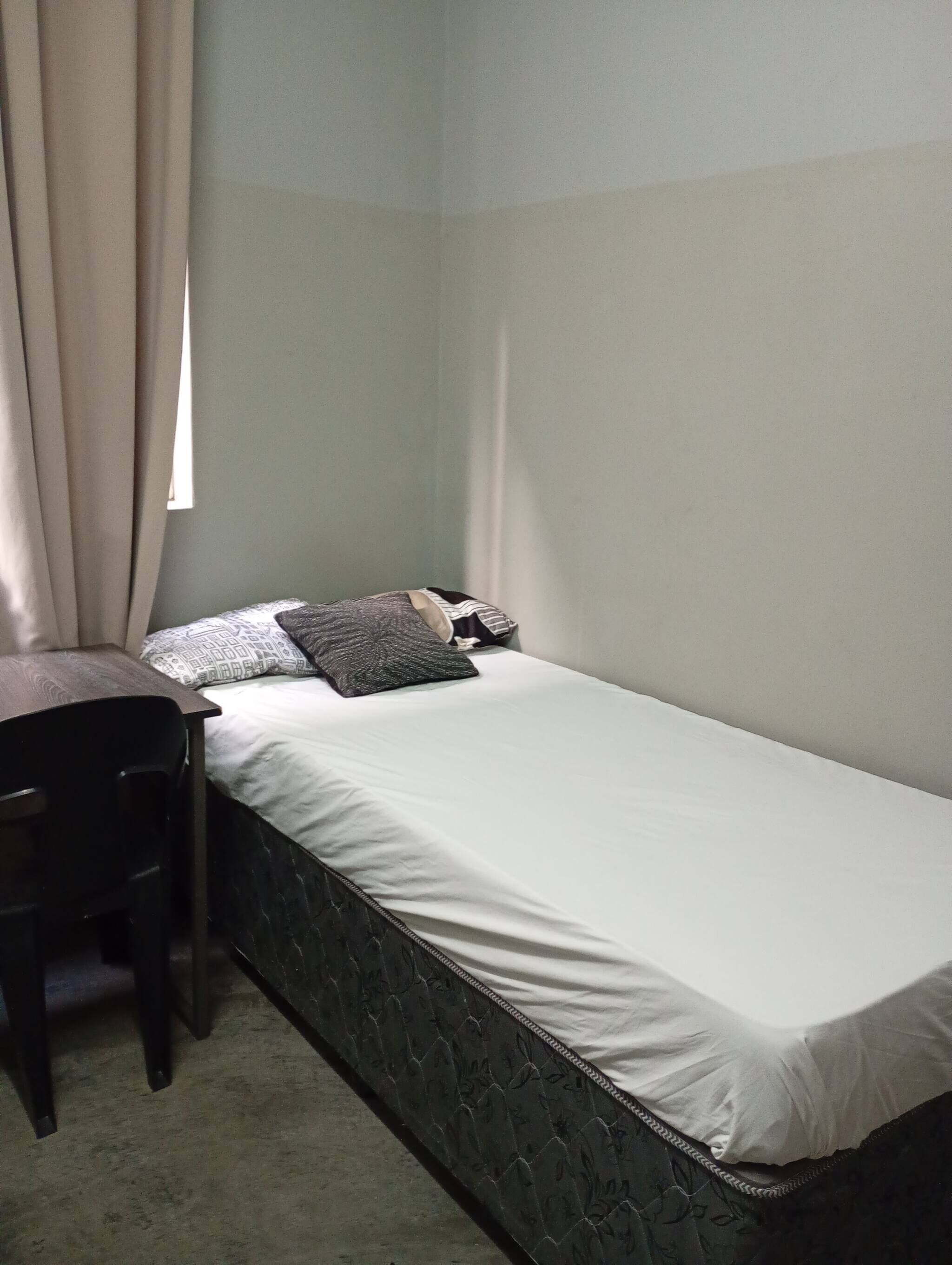 A small, single-bed room with a desk and chair, a window with curtains, and a plain wall.