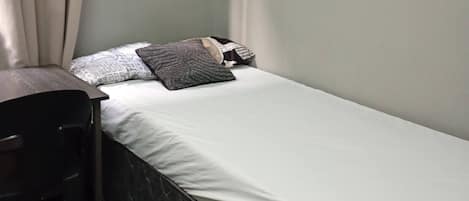 Una habitación pequeña con cama individual, escritorio y silla, ventana con cortinas y una pared sencilla.