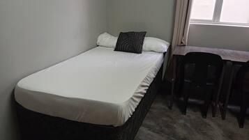 Una cama individual con colchón blanco y cabecera oscura, una mesita con una silla, y una ventana con cortinas.
