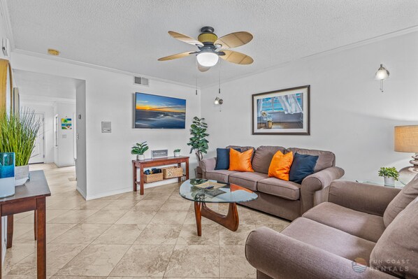 Een moderne woonkamer met een glazen salontafel, een bank met oranje kussens, een plafondventilator en een schilderij van een zonsondergang.
