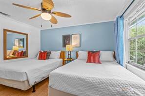 Ein Hotelzimmer mit zwei Betten, einem Deckenventilator, einem großen Fenster mit Vorhängen und Bildern an den Wänden.