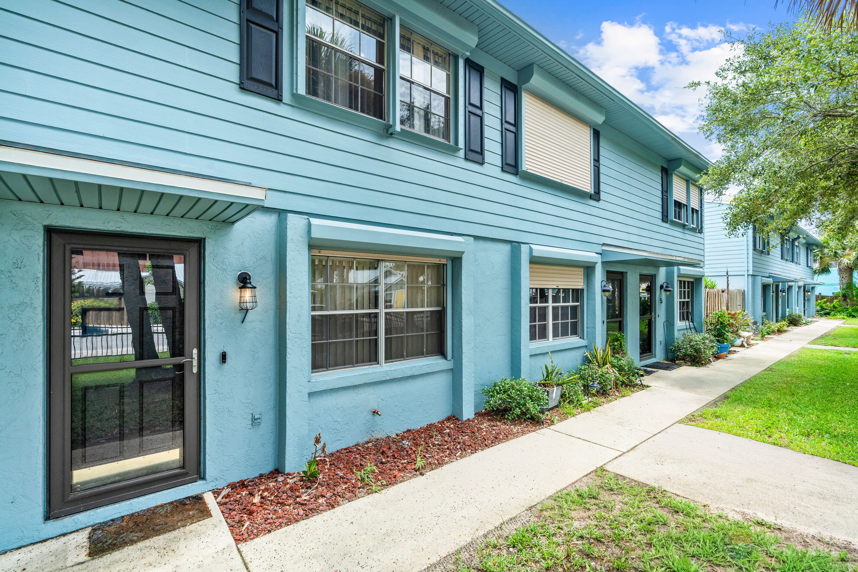 A row of light blue houses with dark shutters, a front door, and a small front yard with mulch and plants.
