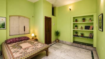 Une chambre à coucher avec un lit, une table de chevet, une lampe, une porte, une étagère avec des plantes et un tableau encadré au mur.
