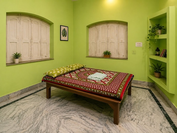 A room with a bed, two windows, a shelf with plants, and a framed picture on the wall.