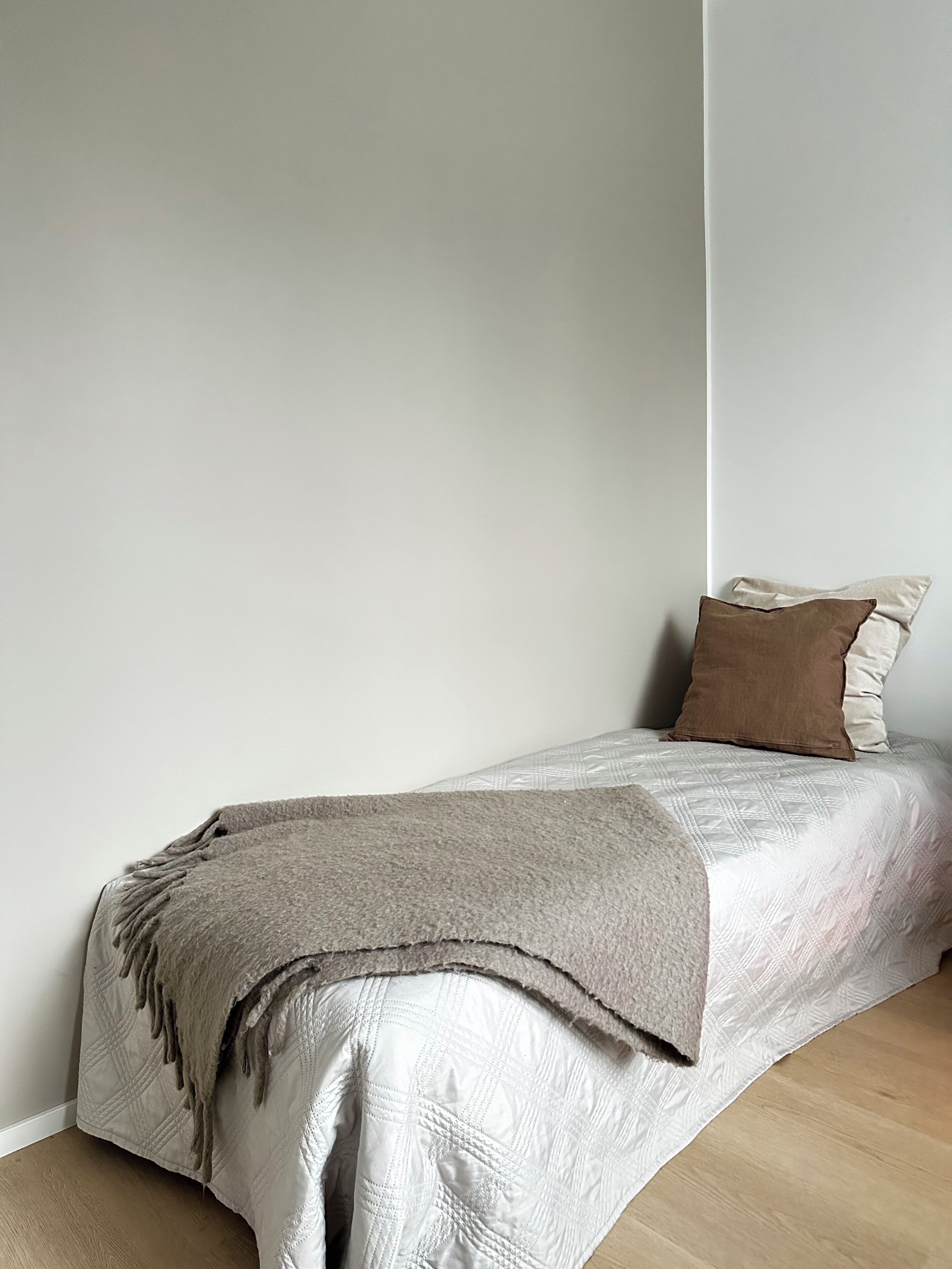 Una cama individual con colcha acolchada y manta, ubicada frente a una pared lisa con suelo de madera.