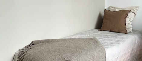 Una cama individual con colcha acolchada y manta, ubicada frente a una pared lisa con suelo de madera.