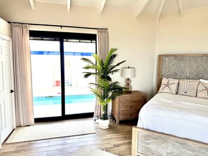 Une chambre avec un lit en bois, une table de chevet avec une lampe, une plante en pot et une vue sur une piscine, visible à travers une porte vitrée.