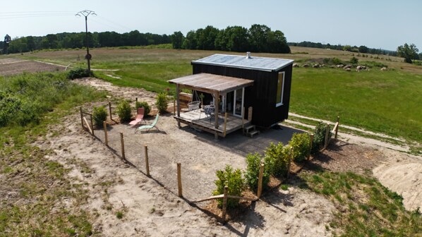Eine kleine, eingeschossige Holzhütte mit Veranda und Sitzbereich im Freien, umgeben von einem eingezäunten Hof und weitläufigen Feldern.
