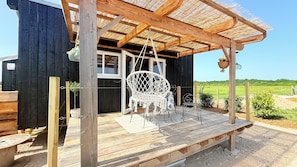 A wooden deck with a hanging chair and a table, a thatched roof, and a view of a green field. - Unique Accommodations (Montrieux-en-Sologne)