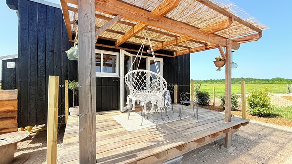 A wooden deck with a hanging chair and a table, a thatched roof, and a view of a green field.