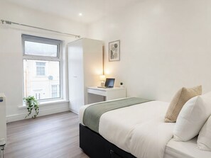 Ein Schlafzimmer mit einem Bett, einem Schreibtisch mit Laptop, einem Fenster und einer Pflanze.