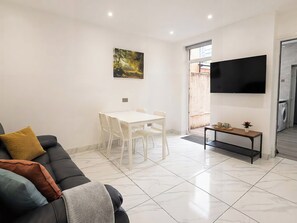Ein modernes Wohnzimmer mit einer Couch, einem Esstisch mit Stühlen, einem flachbildfernseher an der Wand und einem kleinen Beistelltisch mit Tasse und Blume.