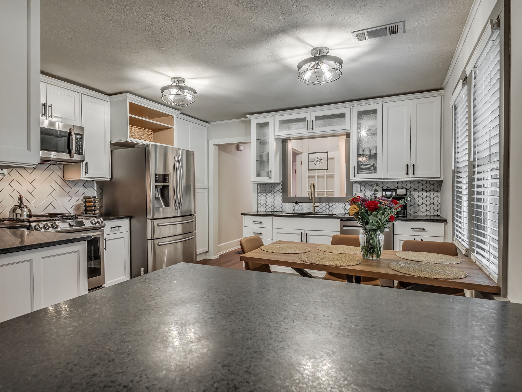 A modern kitchen with stainless steel appliances, a herringbone tile backsplash, and a dining area with a wooden table and woven placemats.