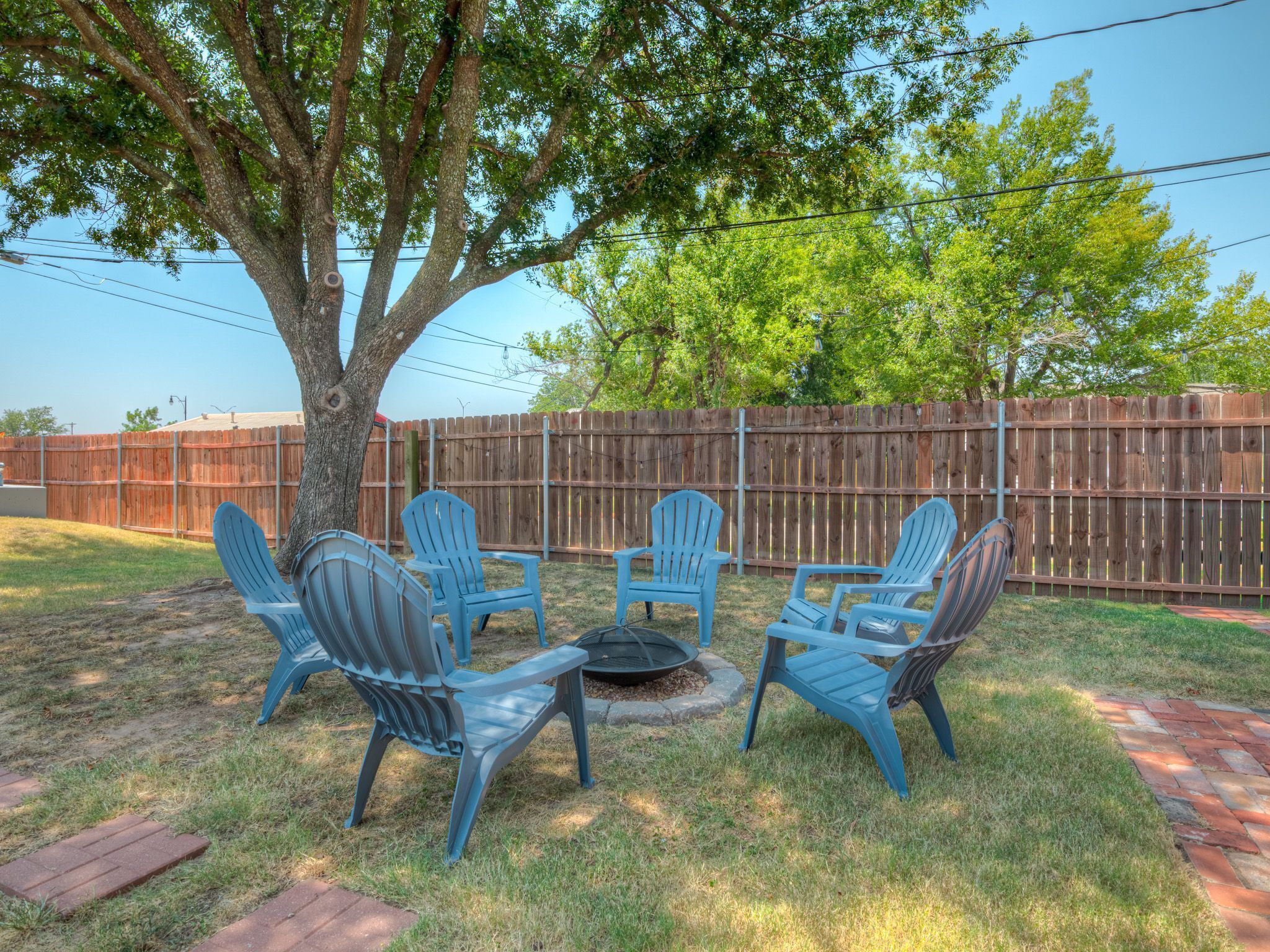 A backyard with a wooden fence, a tree, and a fire pit surrounded by blue chairs.
