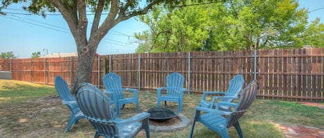 Un patio trasero con cerca de madera, un árbol y un hoyo para fuego rodeado de sillas azules.