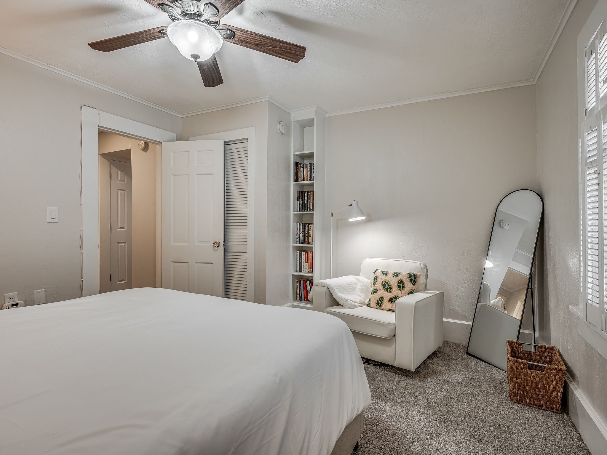 A bedroom with a bed, a white armchair, a bookshelf, a ceiling fan, and a large mirror.