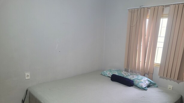 A single bed with a green floral bedspread and a dark blue pillow, a window with beige curtains, and a white wall with a power outlet.