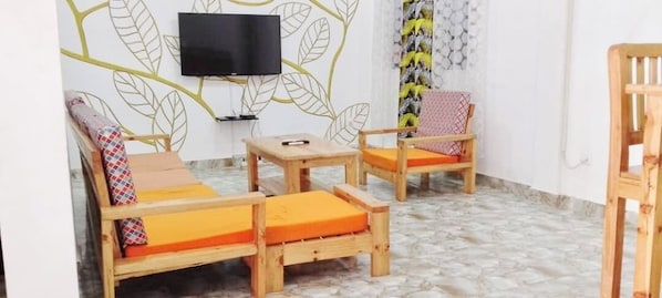 A room with a sofa, two wooden chairs, a small table, and a television mounted on the wall.
