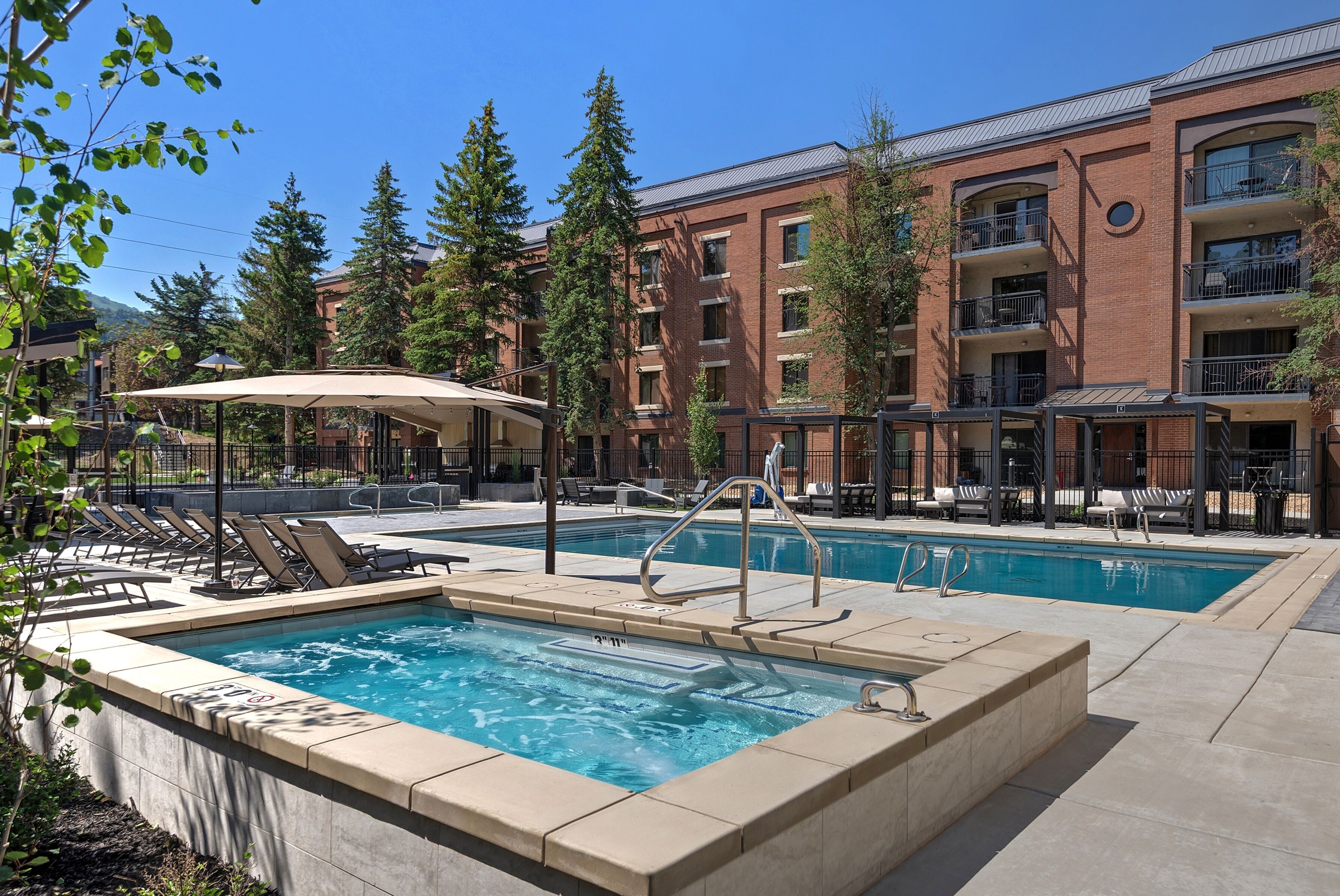 Un espace piscine doté de chaises longues et d’un coin salon abrité, le tout entouré d’un immeuble à plusieurs étages avec des balcons.