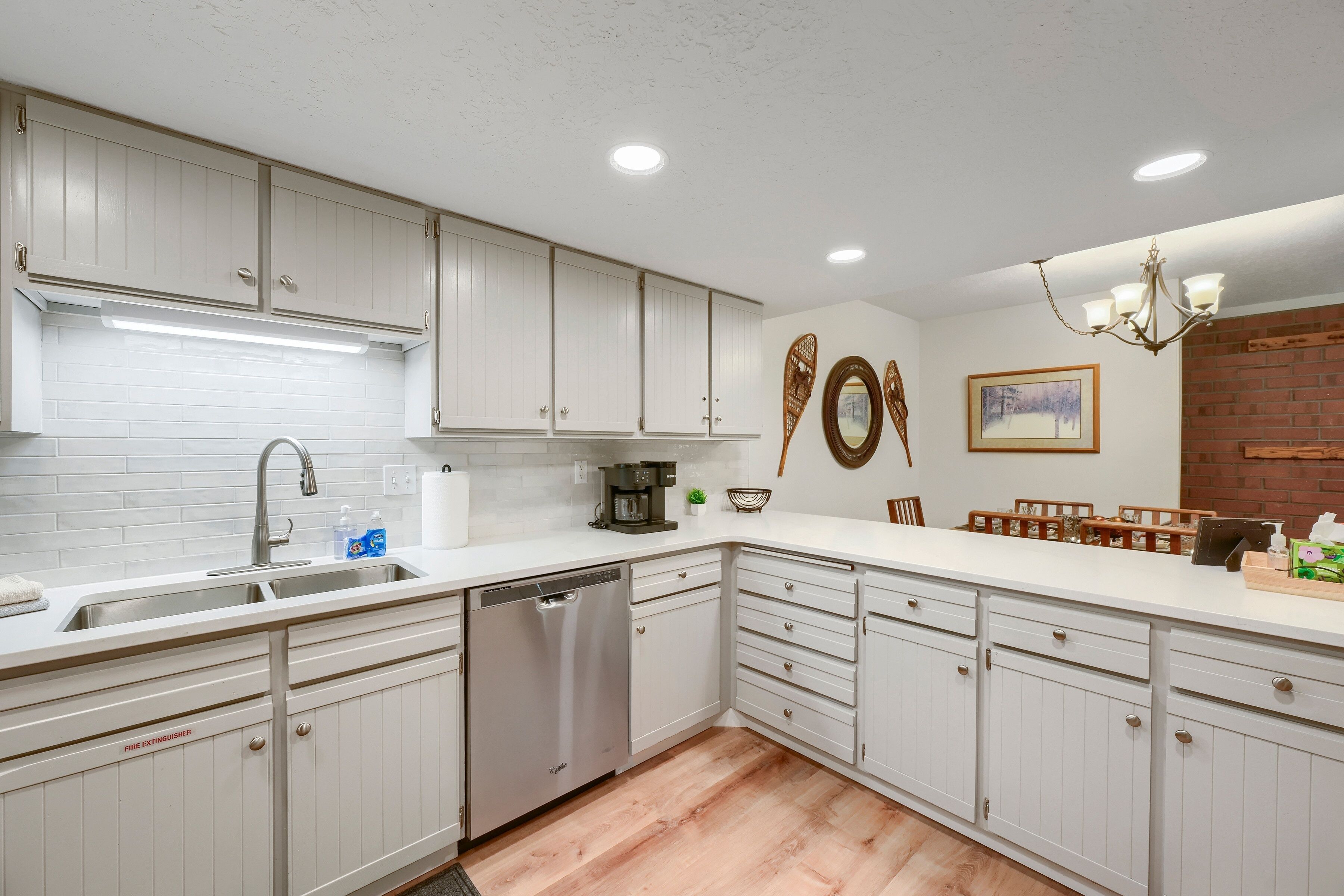 A kitchen with white cabinets, a dishwasher, a coffee maker, and a dining area with a brick wall and a chandelier.