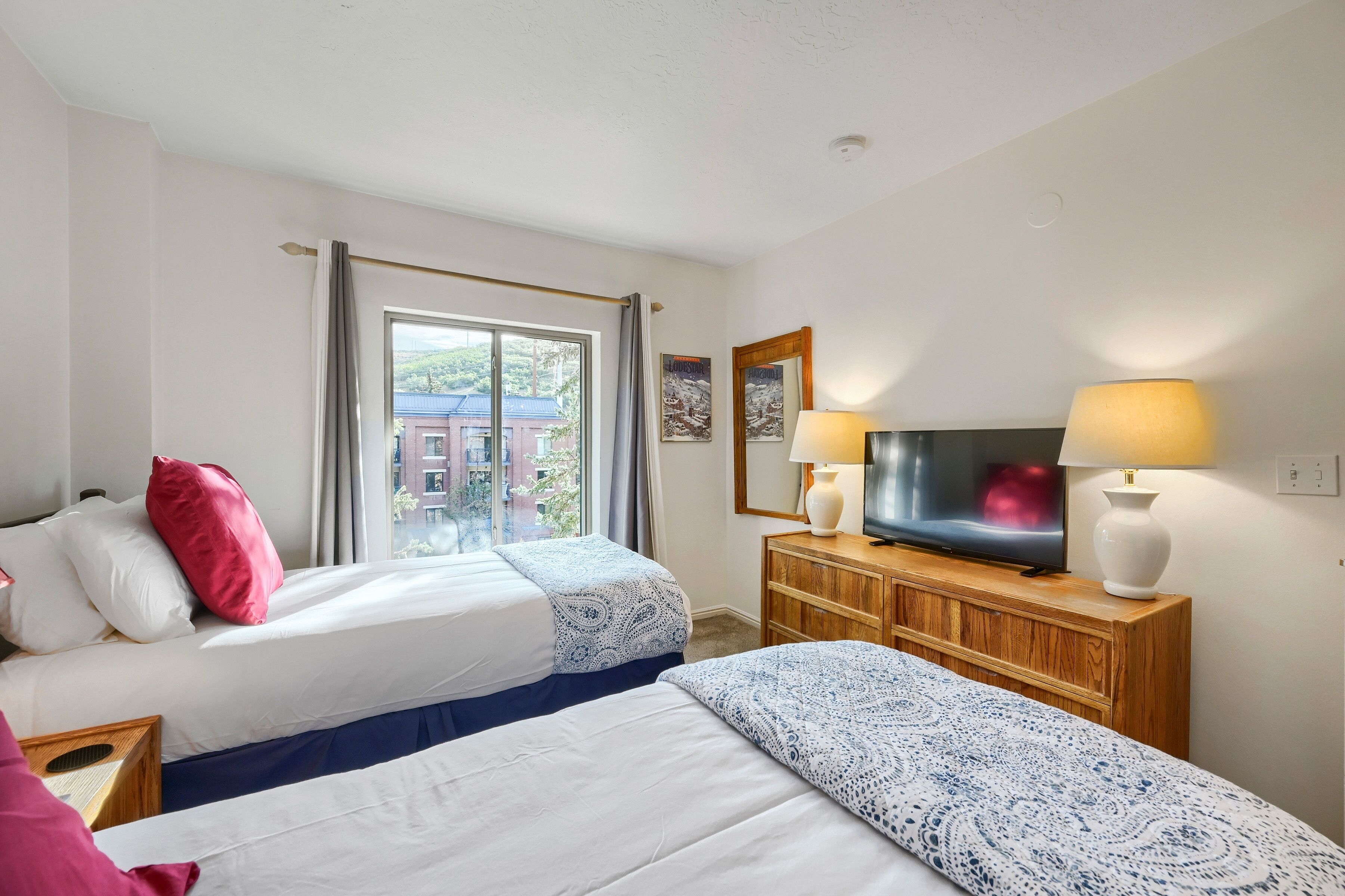 Ein Hotelzimmer mit zwei Betten, einem Fernseher, einer Holzkommode und einem Fenster mit Blick auf Gebäude.