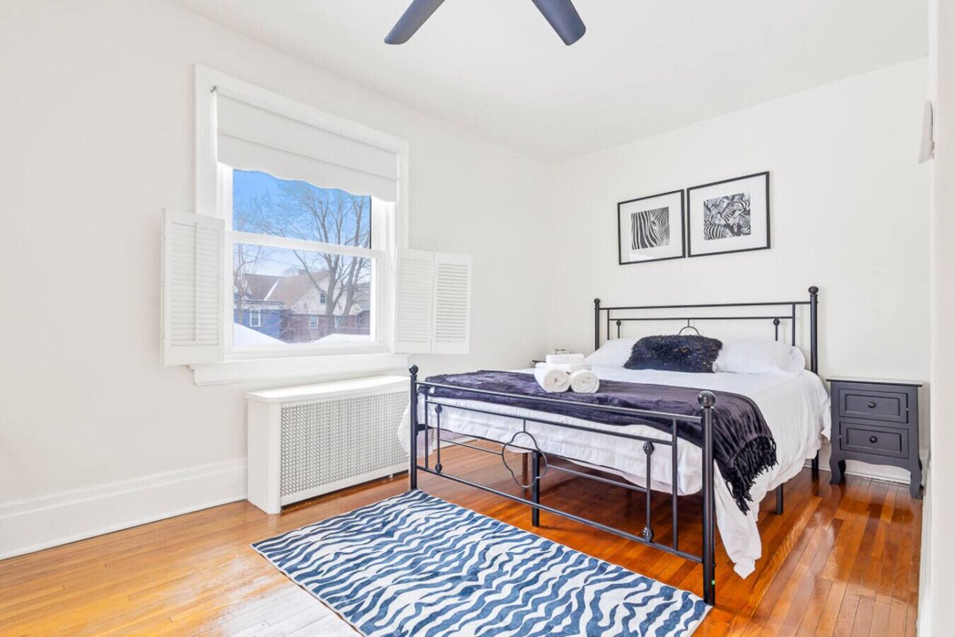 Une chambre à coucher avec un lit, un radiateur, une fenêtre avec des volets et un tapis à motifs zébrés.