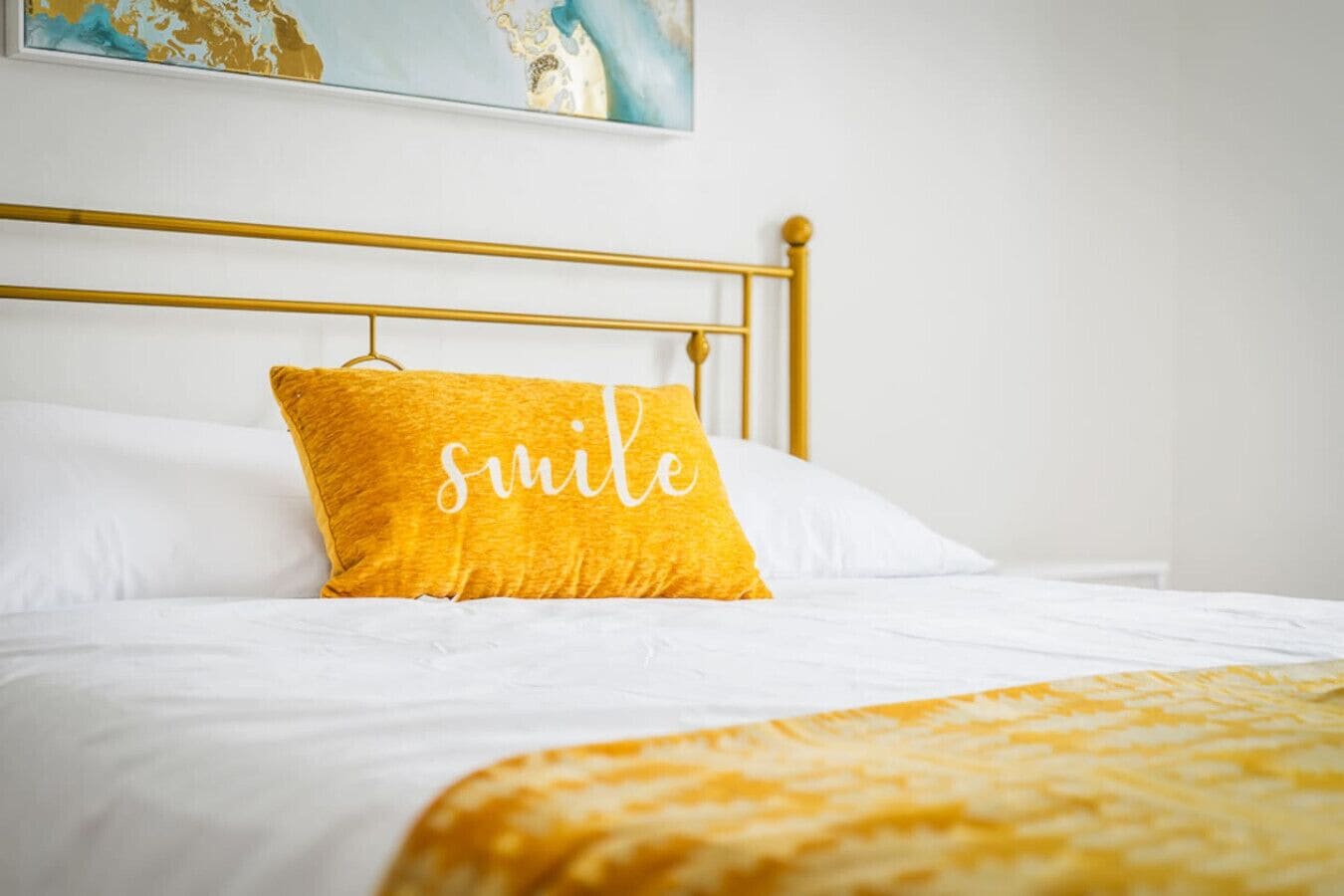 Un lit avec une tête de lit dorée, une literie blanche et un oreiller jaune sur lequel est inscrit le mot « sourire ».