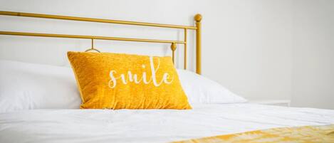 A bed with a gold headboard, white bedding, and a yellow pillow with the word 'smile' on it.