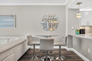 A modern kitchen with a dining area, featuring a glass table, two bar stools, a decorative wall piece, and a pendant light. - Palazzo 1206 Top Destination in PCB!/Oceanfront/+Clean/Wifi/Beach Chairs (Panama City Beach)