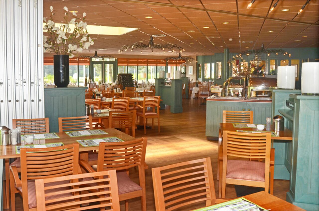 Een restaurant met houten tafels en stoelen, een grote vaas met bloemen en een keuken met een aanrecht en apparatuur.