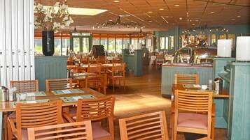 Een restaurant met houten tafels en stoelen, een grote vaas met bloemen en een keuken met een aanrecht en apparatuur.