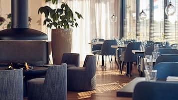 Ein modernes Restaurant-Interieur mit runden Tischen, grau gepolsterten Stühlen, einer großen Küstpflanze und einem Kaminofen.