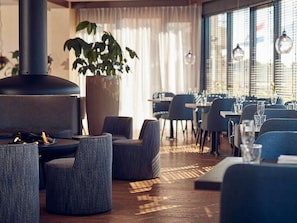 Interior moderno de restaurante con mesas redondas, sillas tapizadas en gris, una planta grande en maceta y un estufa de leña.