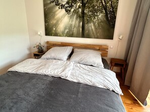 A bedroom with a large bed, a wooden headboard, a nightstand with a lamp, and a wall-mounted painting of a forest. - Apartment on the ski Slope (Bad Kleinkirchheim)