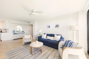 Een moderne woonkamer met een blauwe bank, een witte fauteuil en een klein tafeltje. In de achtergrond is een keuken te zien met witte kasten en een witte plafondventilator.