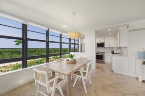 Una cocina moderna con ventanas amplias, un comedor con mesa de madera y sillas de mimbre, y vistas al exterior con vegetación.
